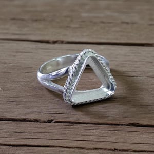 May include: A silver ring with a triangular setting. The ring features a braided design around the triangle and a smooth band. The ring is displayed on a wooden surface.