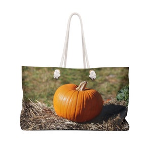 Puede incluir: Una gran calabaza naranja se sienta sobre un lecho de heno. La calabaza está en un campo con hierba verde y una bolsa de lona blanca con asas de cuerda.
