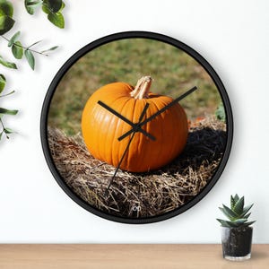 Puede incluir: Un reloj de pared redondo con marco negro y una imagen de una calabaza naranja sentada en un lecho de heno.