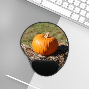 Puede incluir: Una calabaza redonda naranja se sienta en un lecho de heno. La calabaza está enfocada y el heno está desenfocado. La calabaza está sobre una alfombrilla de ratón negra y gris con un círculo negro en el centro.