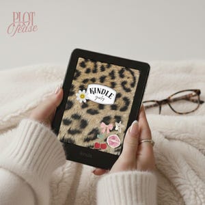 Könnte beinhalten: Ein schwarzer Kindle-E-Reader mit einem Leopardenmuster-Hintergrund, dem Text "Kindle Girly" und dekorativen Aufklebern. Der E-Reader wird vor einem weichen, weißen, strukturierten Hintergrund gehalten.