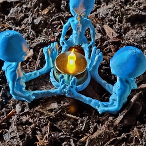 May include: A blue alien-themed candle holder featuring three figures encircling a lit candle. The figures have large heads and slender bodies, holding the candle. The candle holder is set against a backdrop of dark brown mulch.