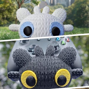 May include: Two plush toys, one white and one dark grey, are positioned above a grey game controller. The white toy has blue eye accents and the dark grey toy has yellow eye accents. The toys have a textured, woven appearance.