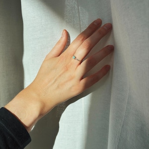 Puede incluir: Un delicado anillo de plata con una pequeña piedra turquesa redonda. El anillo se lleva en una mano, con los dedos ligeramente extendidos. El fondo es una cortina blanca suave y la iluminación es natural.