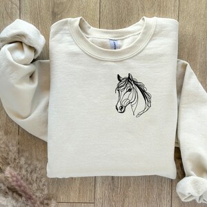 Pu&ograve; includere: Una felpa color crema con un disegno a linea nera di una testa di cavallo sul davanti.