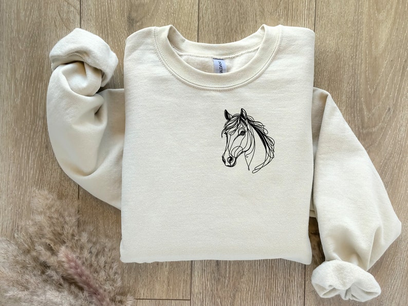 Pu&ograve; includere: Una felpa color crema con un disegno a linea nera di una testa di cavallo sul davanti.