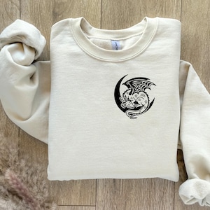 Puede incluir: Una sudadera de color crema con un dise&ntilde;o de drag&oacute;n negro y media luna. El drag&oacute;n est&aacute; detallado con alas y escamas, y est&aacute; centrado en el pecho. La sudadera tiene cuello redondo y mangas largas.