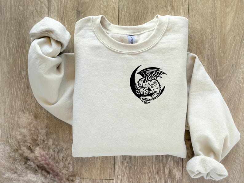 Puede incluir: Una sudadera de color crema con un dise&ntilde;o de drag&oacute;n negro y media luna. El drag&oacute;n est&aacute; detallado con alas y escamas, y est&aacute; centrado en el pecho. La sudadera tiene cuello redondo y mangas largas.