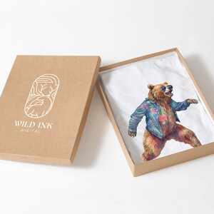 May include: A brown cardboard box with the text "WILD INK DIGITAL" on the front. The box is open and a white t-shirt with a colourful illustration of a bear wearing sunglasses and a floral shirt is inside.