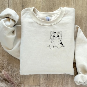 May include: A cream-colored sweatshirt with a black and white cartoon cat peeking out of a pocket on the front.