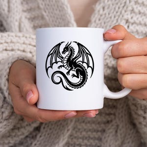 Puede incluir: Taza de cer&aacute;mica blanca con un dise&ntilde;o de drag&oacute;n negro. El drag&oacute;n tiene grandes alas y una larga cola. La taza la sostiene una persona que lleva un jersey color crema. La taza es de tama&ntilde;o est&aacute;ndar.
