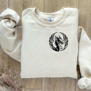 Puede incluir: Sudadera color crema con un dise&ntilde;o de drag&oacute;n negro dentro de un marco circular. El drag&oacute;n est&aacute; representado con alas y una silueta detallada. La sudadera est&aacute; hecha de un material suave y tiene cuello redondo.
