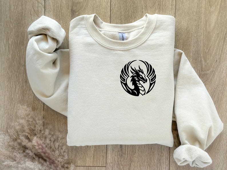 Puede incluir: Sudadera color crema con un dise&ntilde;o de drag&oacute;n negro dentro de un marco circular. El drag&oacute;n est&aacute; representado con alas y una silueta detallada. La sudadera est&aacute; hecha de un material suave y tiene cuello redondo.