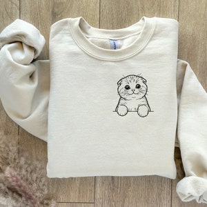 May include: A beige sweatshirt with a black and white illustration of a Scottish Fold cat on the front.