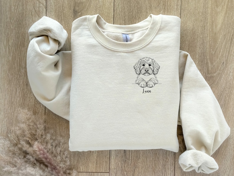 May include: A cream-coloured sweatshirt with a black line drawing of a dog on the front. The dog is looking at the viewer and has the name "Luna" written underneath it.