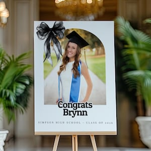 Puede incluir: Un anuncio de graduación en un caballete de madera. El anuncio presenta una foto de una graduada con birrete y toga, con el texto "Congrats Brynn" y "Simpson High School - Class of 2016". Un lazo negro en la parte superior.