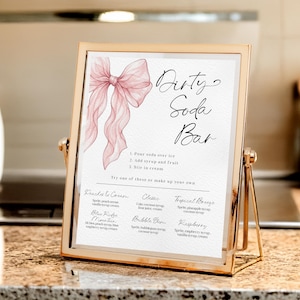May include: A gold-framed sign with the words "Dirty Soda Bar" in elegant script. The sign features a watercolor illustration of a pink bow and a list of soda recipes. The sign is displayed on a countertop.