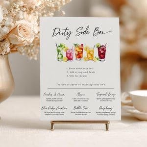 May include: A white sign with the text "Dirty Soda Bar" and instructions for making soda. The sign features watercolor illustrations of colorful soda drinks and a gold stand. The sign also lists soda recipes.