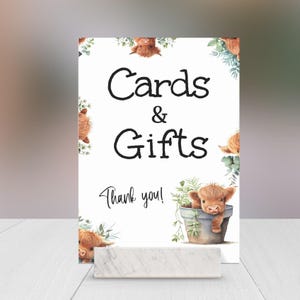 May include: White sign with black text that reads "Cards & Gifts Thank you!" with a border of cartoon highland cows peeking out from behind green foliage.