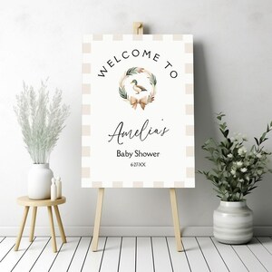 May include: Welcome to Amelia's Baby Shower sign with a white and beige gingham background. A wreath with a duck and a bow is in the center. The text "Welcome to Amelia's Baby Shower 6-27-XX" is printed on the sign.