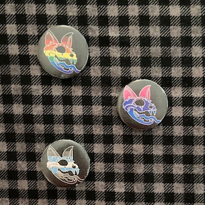 May include: Three round black and white buttons with a skull and crossbones design. Each button features a different color scheme: rainbow, bisexual, and transgender.