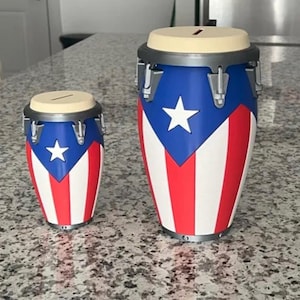 May include: Two conga drum-shaped money banks. The drums are painted with the Puerto Rican flag design: blue with a white star, and red and white stripes. The tops are beige with a slot for inserting money.