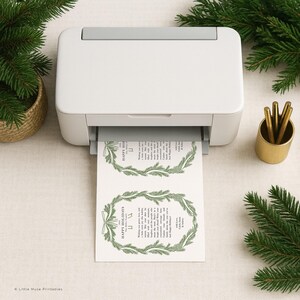 K&ouml;nnte beinhalten: Ein wei&szlig;er Drucker druckt zwei Weihnachtsgru&szlig;karten mit gr&uuml;nen Kranzdesigns und dem Text "Happy Holidays". Die Karten liegen auf einer neutralen Oberfl&auml;che, mit einem goldenen Stifthalter und Tannenzweigen.