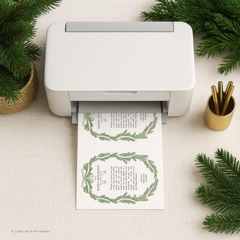 K&ouml;nnte beinhalten: Ein wei&szlig;er Drucker druckt zwei Weihnachtsgru&szlig;karten mit gr&uuml;nen Kranzdesigns und dem Text "Happy Holidays". Die Karten liegen auf einer neutralen Oberfl&auml;che, mit einem goldenen Stifthalter und Tannenzweigen.