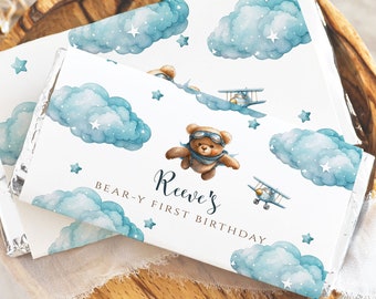 May include: White chocolate bar wrapper with a blue and white watercolor design of clouds, stars, and a teddy bear wearing aviator goggles and flying a plane. The text "Reeve's Bear-y First Birthday" is printed on the wrapper.