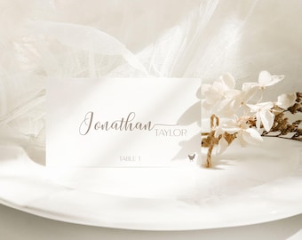 May include: A white place card with the name "Jonathan" in elegant script, followed by "Taylor" in a smaller font. The card also includes "Table 1" and a small chicken graphic. The card is set on a white plate, with white flowers and a white fabric in the background.