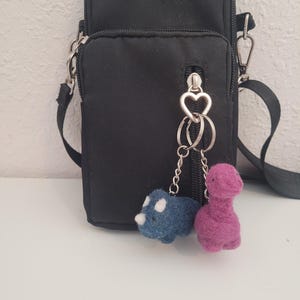 May include: Black crossbody bag with a zippered front pocket and a silver-tone heart-shaped zipper pull. The bag has a black adjustable strap. Attached to the zipper pull are two small felt dinosaur keychains, one blue with white spots and one purple.