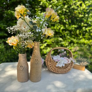 Könnte beinhalten: Zwei braune Glasflaschen, die mit Schnur umwickelt sind und Holzhertz-Akzente haben, halten gelbe und weiße Blumen. Ein Weidenkorb mit rosa Blumen und ein Holzschild mit der Aufschrift "Drei" sind ebenfalls im Bild.