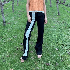 May include: A person wearing a peach-colored, short-sleeved top with a chevron pattern and black pants with a white stripe down the side. The person is standing in a vineyard with green grass and bare vines.