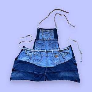 Könnte beinhalten: Eine mehrfarbige blaue Denim-Schürze aus wiederverwerteten Jeans. Das Design umfasst ein Latzteil mit einer mittigen Tasche, einen Taillenbereich mit zwei Gesäßtaschen und ein breites Unterteil. Helle Stoffbänder sind am Hals und an der Taille befestigt.