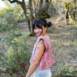 Könnte beinhalten: Eine Person trägt ein pinkfarbenes und lilafarbenes, gestreiftes, bauchfreies Oberteil mit einem schwarz-weißen grafischen Druck auf dem Arm. Die Person trägt Jeans-Shorts und steht in einem Waldgebiet.