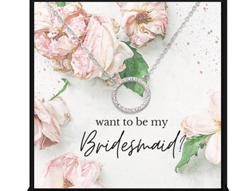 May include: A white box with a floral design and the text "want to be my Bridesmaid?"  A silver necklace with a circle pendant is displayed on top of the box.