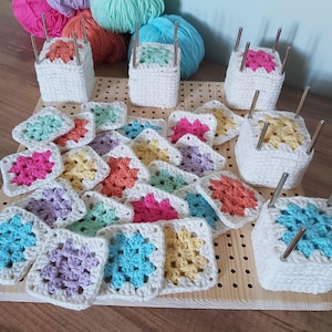 Op de afbeelding: Een houten opspanbord met een raster van gaten, voorzien van verschillende witte gehaakte vierkante blokken met metalen pinnen en een verzameling veelkleurige gehaakte granny squares in roze, gele, blauwe en paarse tinten op het oppervlak.