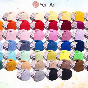 Op de afbeelding: Een verzameling kleurrijke bollen garen in diverse tinten, waaronder wit, oranje, rood, roze, paars, blauw, groen, geel en bruin. Elke bol heeft een label met het "YarnArt"-logo. Het garen is in nette rijen gerangschikt.