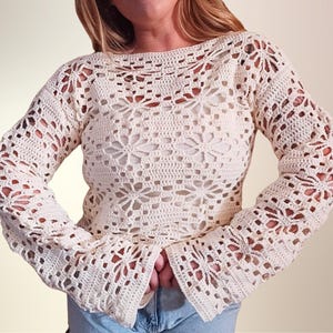 May include: A long-sleeved, off-white crochet sweater featuring an intricate, repeating floral pattern throughout the garment. The design includes a wide, open-knit structure with a boat neckline and flared sleeves, crafted from a light-colored yarn.