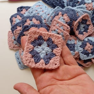 Può includere: Una collezione di piccoli motivi quadrati all'uncinetto con un centro azzurro, circondato da uno strato blu scuro e rifinito con un bordo rosa polvere. Questi quadrati della nonna fatti a mano sono impilati in un mucchio.