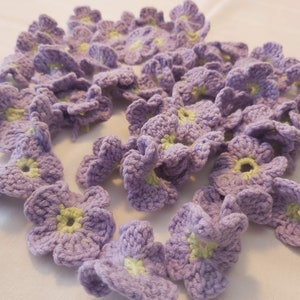 May include: A collection of crocheted lavender flowers with light green centres. The flowers are arranged in a pile on a white surface.