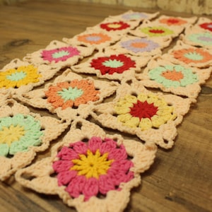 May include: A rectangular crochet table runner composed of multiple square motifs joined together. Each square features a central flower design in various colors including red, yellow, pink, orange, and light green, all framed by a light cream yarn border.