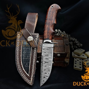 May include: A Damascus steel hunting knife with a wooden handle and a leather sheath. The knife is resting on a wooden box with dominoes scattered around it. The text "Duck n Deer" is visible in the bottom right corner of the image.