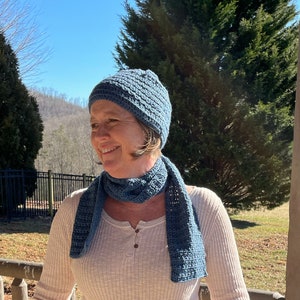 Peut inclure: Une femme portant un bonnet et une écharpe en crochet bleu. Le bonnet et l'écharpe sont en point texturé. La femme porte une chemise blanche à manches longues.