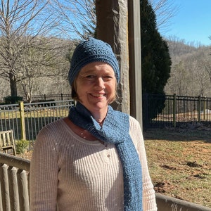 May include: A woman wearing a blue knitted hat and a blue knitted scarf. She is standing in front of a wooden railing and a green grassy area.