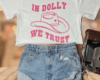 Può includere: Maglietta bianca corta con testo rosa che recita "In Dolly We Trust" e un'immagine di un cappello da cowboy rosa.