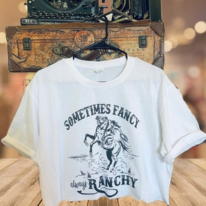 Op de afbeelding: Wit gecropped T-shirt met een zwarte afbeelding van een cowboy die een bokkend paard berijdt. De tekst "Sometimes Fancy always Ranch" is op het shirt gedrukt.