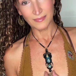 May include: A black macrame necklace featuring a large, dark teal oval gemstone and a smaller matching stone, accented with gold-toned metal beads and a spiral charm. The necklace is worn with gold-toned filigree hoop earrings and a mustard-colored top.