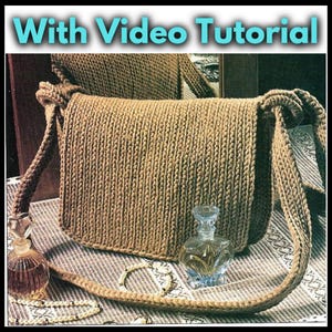Op de afbeelding: Een bruine gehaakte schoudertas met een lange band. De tas ligt op een tafel met een wit kantkleed. De tekst "With Video Tutorial" staat bovenaan de afbeelding.