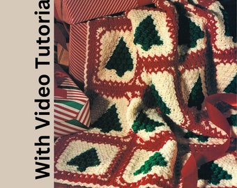May include: A handmade crochet blanket with a Christmas tree design. The blanket features green trees on a cream background, framed by red borders. The text "With Video Tutorial" is on the left side of the image. Red ribbon and wrapped gifts are in the background.